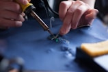 A person is soldering electronic components onto a circuit board using a soldering iron. The scene includes detailed view of the hands holding the soldering iron and the circuit board, with visible smoke rising from the solder.