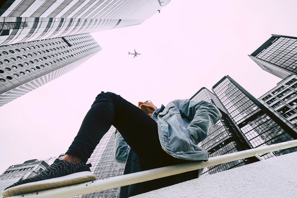 low-angle photography of person wearing blue denim jacket and black pants leaning on white metal railing between curtain wall buildings during daytime