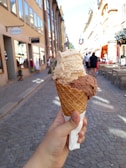 A hand holds an ice cream cone with two scoops of ice cream, one beige and one chocolate brown. The cone has a waffle texture and is wrapped at the bottom with a napkin. The background features a cobblestone pedestrian street with storefronts on either side under a clear blue sky. People walk along the street, which has outdoor seating available.