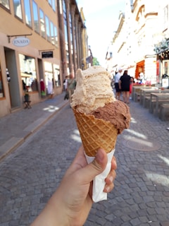 A hand holds an ice cream cone with two scoops of ice cream, one beige and one chocolate brown. The cone has a waffle texture and is wrapped at the bottom with a napkin. The background features a cobblestone pedestrian street with storefronts on either side under a clear blue sky. People walk along the street, which has outdoor seating available.