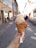 A hand holds an ice cream cone with two scoops of ice cream, one beige and one chocolate brown. The cone has a waffle texture and is wrapped at the bottom with a napkin. The background features a cobblestone pedestrian street with storefronts on either side under a clear blue sky. People walk along the street, which has outdoor seating available.