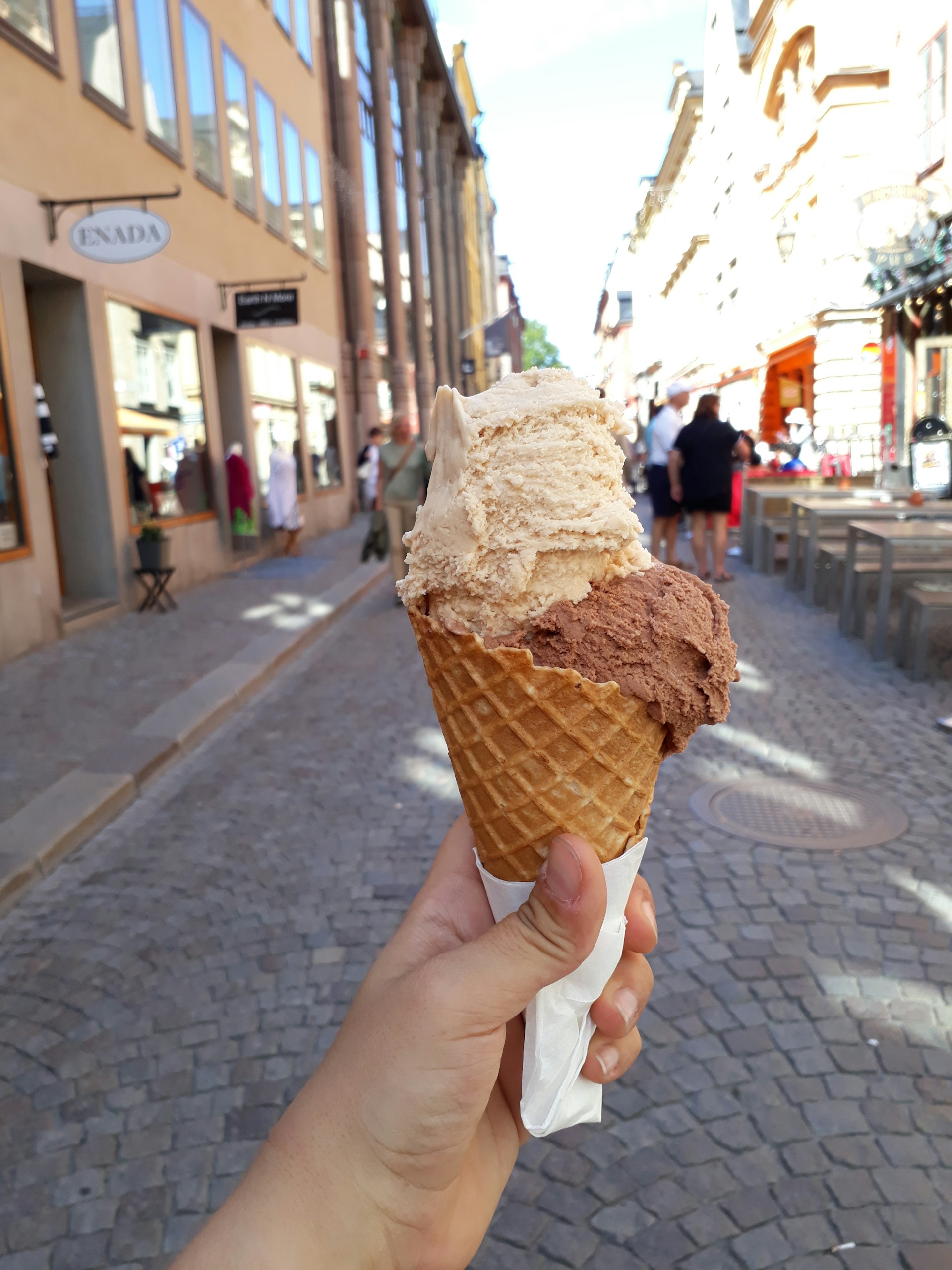 A hand holds an ice cream cone with two scoops of ice cream, one beige and one chocolate brown. The cone has a waffle texture and is wrapped at the bottom with a napkin. The background features a cobblestone pedestrian street with storefronts on either side under a clear blue sky. People walk along the street, which has outdoor seating available.