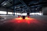 A drone is positioned on the floor of a large, dimly lit industrial warehouse. Its lights are glowing red, casting a warm glow on the concrete surface. The vast space features metal beams and scattered debris, with light filtering through high windows.