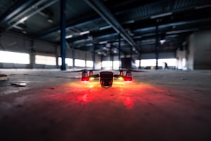A drone is positioned on the floor of a large, dimly lit industrial warehouse. Its lights are glowing red, casting a warm glow on the concrete surface. The vast space features metal beams and scattered debris, with light filtering through high windows.