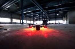 A drone is positioned on the floor of a large, dimly lit industrial warehouse. Its lights are glowing red, casting a warm glow on the concrete surface. The vast space features metal beams and scattered debris, with light filtering through high windows.