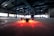 A drone is positioned on the floor of a large, dimly lit industrial warehouse. Its lights are glowing red, casting a warm glow on the concrete surface. The vast space features metal beams and scattered debris, with light filtering through high windows.