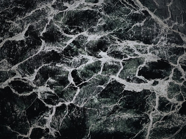 The image displays an intricate pattern of dark green and black marble with white veining. The texture appears smooth and polished, with the veins creating a natural, abstract design across the surface.