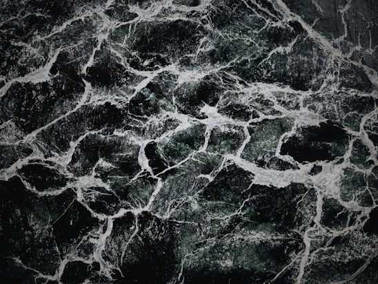 The image displays an intricate pattern of dark green and black marble with white veining. The texture appears smooth and polished, with the veins creating a natural, abstract design across the surface.