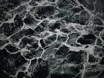 The image displays an intricate pattern of dark green and black marble with white veining. The texture appears smooth and polished, with the veins creating a natural, abstract design across the surface.