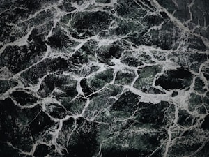 The image displays an intricate pattern of dark green and black marble with white veining. The texture appears smooth and polished, with the veins creating a natural, abstract design across the surface.