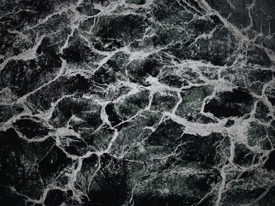 The image displays an intricate pattern of dark green and black marble with white veining. The texture appears smooth and polished, with the veins creating a natural, abstract design across the surface.