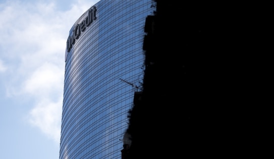 A modern glass skyscraper with reflective windows and a visible company logo near the top. Part of the building is cast in shadow, highlighting the contrast between the illuminated and darker areas. The sky is partly cloudy.