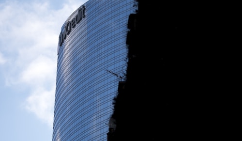 A modern glass skyscraper with reflective windows and a visible company logo near the top. Part of the building is cast in shadow, highlighting the contrast between the illuminated and darker areas. The sky is partly cloudy.