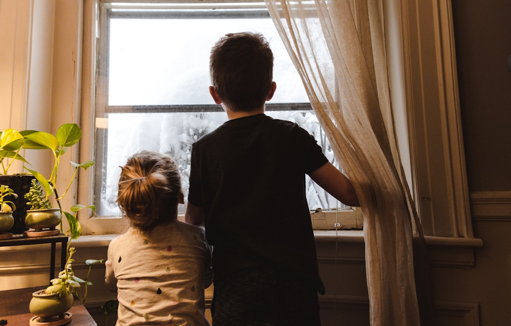 niño y niña de pie cerca de la ventana mirando hacia afuera