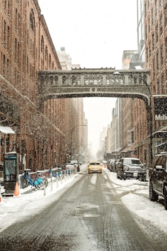 A snowy urban scene featuring a narrow street lined with tall brick buildings on both sides. A historic-looking iron bridge spans the street above. A yellow taxi is driving down the slushy road, surrounded by parked cars. Snow is actively falling, and a row of bicycles is visible on a rack along the sidewalk.