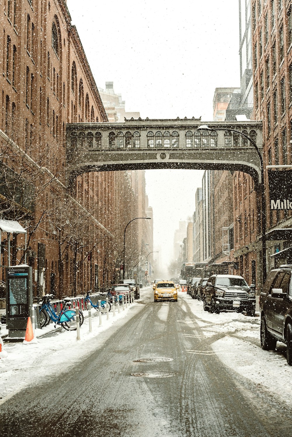 veículo amarelo entre prédios marrons além de veículos estacionados enquanto chove neve