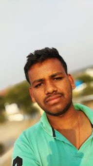 A young man with short dark hair is looking directly at the camera. He is wearing a light green polo shirt with a small number printed on the sleeve. There appears to be a red thread around his neck. The background is slightly blurred, with trees and possibly some buildings visible.