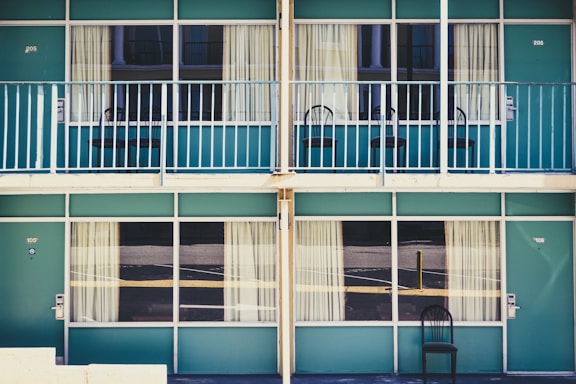 A symmetrical view of a retro-style motel with teal doors and railings. There are two floors, each with large windows draped in beige curtains, and single black chairs placed outside each room. The design appears minimal, with no people present.