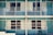 A symmetrical view of a retro-style motel with teal doors and railings. There are two floors, each with large windows draped in beige curtains, and single black chairs placed outside each room. The design appears minimal, with no people present.