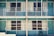 A symmetrical view of a retro-style motel with teal doors and railings. There are two floors, each with large windows draped in beige curtains, and single black chairs placed outside each room. The design appears minimal, with no people present.