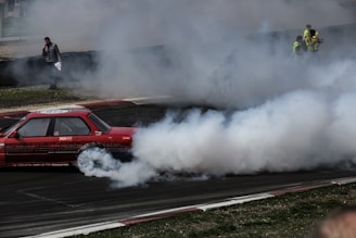 A red car is drifting on a racetrack, creating a large cloud of smoke behind it. Several people in fluorescent vests are observing the scene from the sidelines, while one person stands closer to the track holding papers.