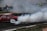 A red car is drifting on a racetrack, creating a large cloud of smoke behind it. Several people in fluorescent vests are observing the scene from the sidelines, while one person stands closer to the track holding papers.