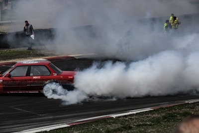 A red car is drifting on a racetrack, creating a large cloud of smoke behind it. Several people in fluorescent vests are observing the scene from the sidelines, while one person stands closer to the track holding papers.