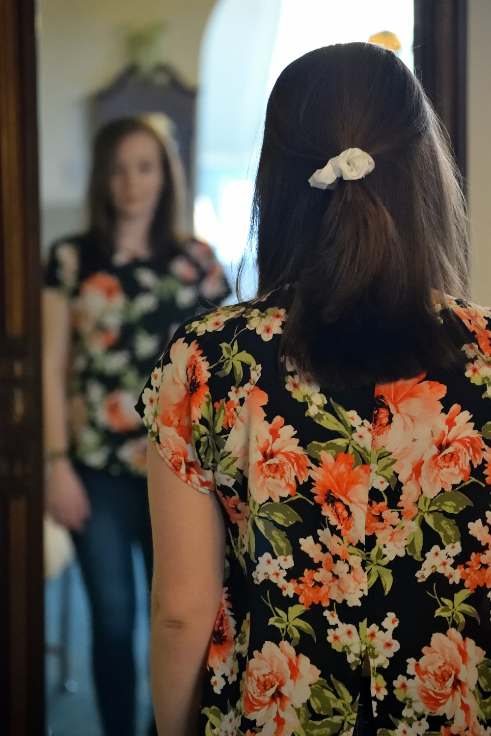 Femme portant un chemisier noir et multicolore et un jean en denim bleu face au miroir à l’intérieur de la chambre en béton blanc