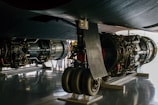 A close-up view of the undercarriage of an aircraft, showcasing large jet engines and landing gear. The engines are detailed with visible mechanical components, and the scene is well-lit with a reflective surface on the floor. Some people are visible in the background, slightly out of focus.