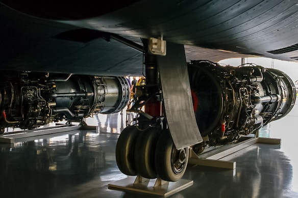 A close-up view of the undercarriage of an aircraft, showcasing large jet engines and landing gear. The engines are detailed with visible mechanical components, and the scene is well-lit with a reflective surface on the floor. Some people are visible in the background, slightly out of focus.