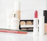 An arrangement of various cosmetic products including a tube of pink lipstick, an eyeshadow palette, lip liners, foundation, and skincare products. The items are placed against a soft, blurred white background, emphasizing the products.