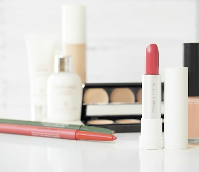 An arrangement of various cosmetic products including a tube of pink lipstick, an eyeshadow palette, lip liners, foundation, and skincare products. The items are placed against a soft, blurred white background, emphasizing the products.