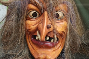 A detailed wooden mask of a witch with exaggerated features, including a large crooked nose, wide eyes, and large teeth. The mask is adorned with long, wild brown hair.