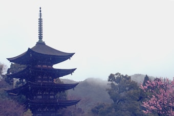 A traditional multi-tiered pagoda stands prominently surrounded by lush green trees, with pink cherry blossoms in bloom nearby. Mist envelops the distant hills, adding a serene and tranquil atmosphere.