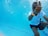 A person underwater in a swimming pool giving a thumbs-up gesture, bubbles surrounding them in clear blue water. The person is wearing a white shirt and dark shorts with an orange wristband.