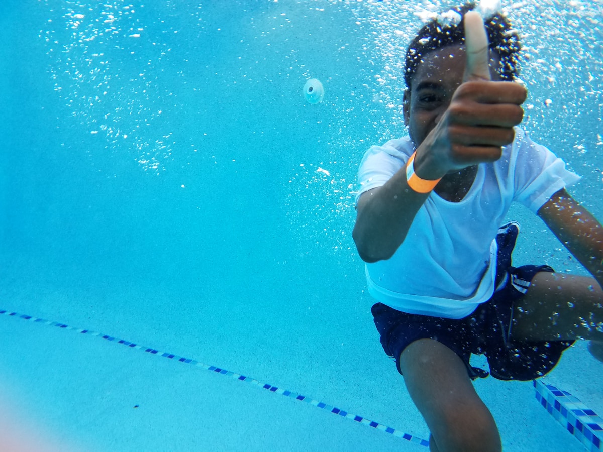 A little brown skinned boy with short hair wearing a white swim shirt and blue trunks with two white stripes underwater in a pool.  He is surrounded by tiny bubbles and has his right hand outstretched with a thumbs up.