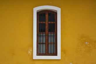 A wooden window with iron bars is set within a white frame, positioned on a vivid yellow wall. The window has a vertical glass pane divided into four sections by wooden bars. The wall shows signs of wear with small patches where the paint has chipped off.
