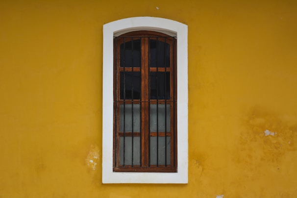A wooden window with iron bars is set within a white frame, positioned on a vivid yellow wall. The window has a vertical glass pane divided into four sections by wooden bars. The wall shows signs of wear with small patches where the paint has chipped off.