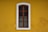A wooden window with iron bars is set within a white frame, positioned on a vivid yellow wall. The window has a vertical glass pane divided into four sections by wooden bars. The wall shows signs of wear with small patches where the paint has chipped off.