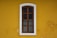 A wooden window with iron bars is set within a white frame, positioned on a vivid yellow wall. The window has a vertical glass pane divided into four sections by wooden bars. The wall shows signs of wear with small patches where the paint has chipped off.
