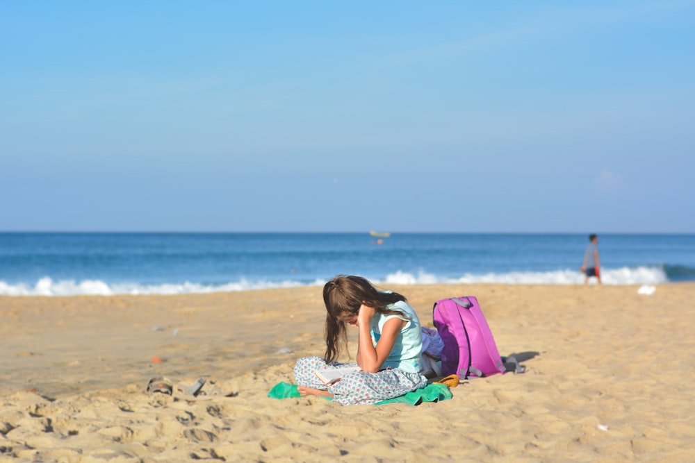 소녀는 푸른 하늘 아래 보라색 배낭 근처 해안에 앉아 있습니다.