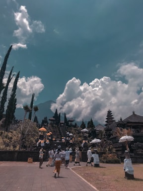 A group of people dressed in traditional attire are walking towards a temple complex with tall, tiered pagoda-style roofs. The sky above is filled with large, fluffy clouds and a mountain silhouette can be seen in the background. Tall trees line the pathway leading to the temple.