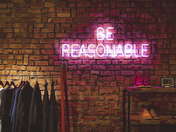 A neon sign displaying the words 'BE REASONABLE' is mounted on an exposed brick wall. Below the sign, clothing on hangers is visible on the left, and on the right, there is a shelf displaying shoes and a framed picture.