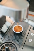 Espresso is being prepared in a metal cup under a professional espresso machine. The coffee has a rich crema on top, and the equipment features a modern, stainless steel design with perforations for drainage.