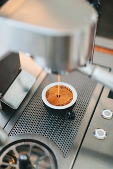 Espresso is being prepared in a metal cup under a professional espresso machine. The coffee has a rich crema on top, and the equipment features a modern, stainless steel design with perforations for drainage.