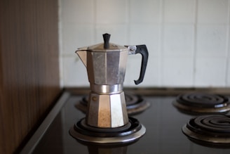 A stovetop espresso maker, often called a moka pot, sits on an electric coil burner. The silver metal contrasts against the dark burners and the tiled backsplash, with the black handle and top knob adding a touch of contrast. The setting suggests a kitchen environment.