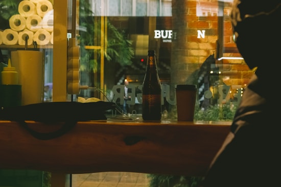 A cozy cafe scene with a wooden counter featuring a beer bottle and a takeaway coffee cup. The background shows a brick wall and a window with reflections of outside greenery and urban elements. Several paper towel rolls and condiment bottles are positioned on the left.