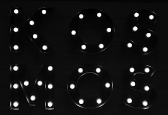Illuminated letters form the word 'KOSMOS'. Each letter is decorated with individual bright bulbs against a dark background, creating a striking contrast.