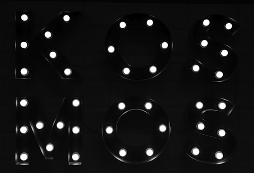 Illuminated letters form the word 'KOSMOS'. Each letter is decorated with individual bright bulbs against a dark background, creating a striking contrast.