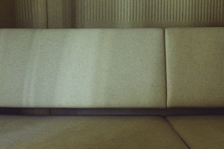 A close-up view of a minimalist sofa with a textured fabric finish. The upholstery features a subtle olive green tone, set against a backdrop of matching tones. The fabric appears softly lit, highlighting its natural texture.